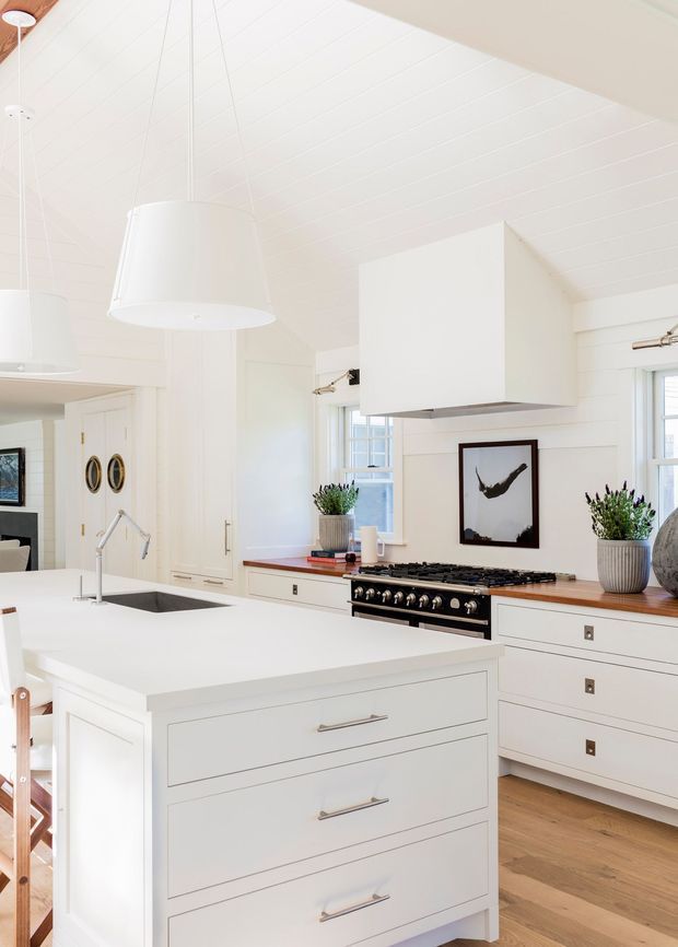 Bright white kitchen with a large central island, integrated sink and gooseneck tap, extensive white cabinetry, a professional black range cooker, timber benchtops, white subway-style wall cladding, and light timber flooring. The room features a white pitched or vaulted panelled ceiling, pendant lighting, large windows, and restrained coastal farmhouse detailing.
