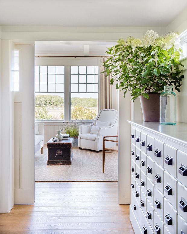 Bright, refined interior view through a wide doorway into a sitting room. The room features large black-grid windows, white-painted timber wall panelling and trim, pale timber flooring, a light upholstered armchair, a small dark wood chest table, and a large white built-in drawer unit in the foreground topped with leafy flowering branches and a glass vessel. The palette is predominantly white and light wood with abundant natural light and a calm, classic feel.