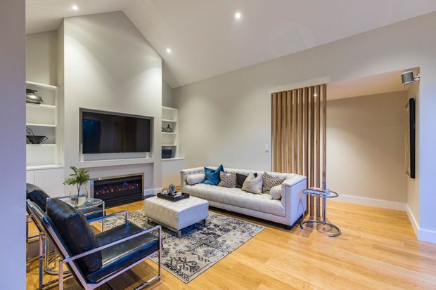 Contemporary living room with a recessed wall-mounted television, built-in open shelving, and a linear fireplace set within pale built-in joinery. A light upholstered sofa, black leather lounge chairs, patterned area rug, glass coffee table, and side tables occupy the room. Vertical timber slats screen an opening or doorway, while recessed ceiling lights and pale walls create a bright, refined interior.