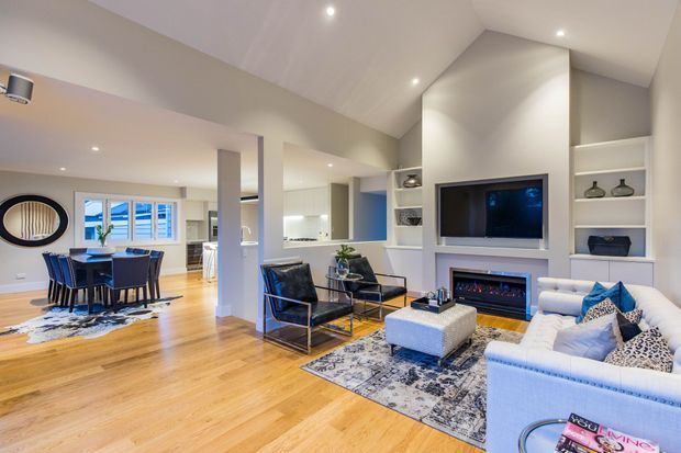 Bright open-plan contemporary living and dining area with warm timber flooring, white walls and built-in joinery. The living room features a large sectional sofa, black leather occasional chairs, patterned rug, wall-mounted television and a built-in fireplace. A dining table and kitchen are visible in the adjoining open space, with recessed lighting and shelving throughout.