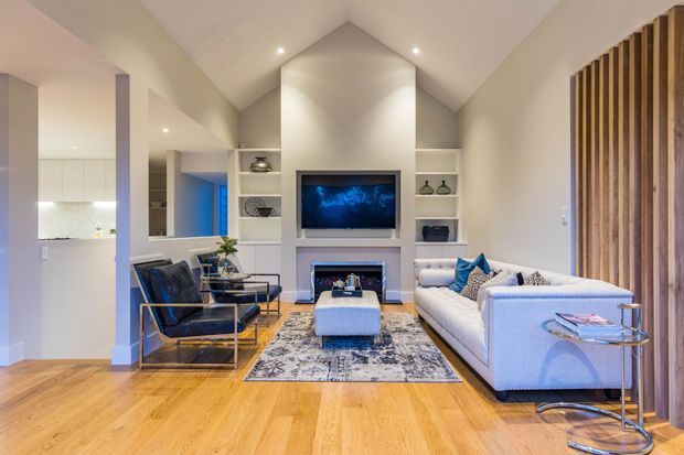 A bright, contemporary open-plan living room with a vaulted ceiling, warm timber flooring, white built-in shelving and fireplace cabinetry, and a wall-mounted television. The room includes a large light-coloured sofa, black leather armchairs, a central ottoman, patterned rug, glass side tables, and a striking vertical timber-slat feature wall. A kitchen area is visible through the open-plan layout.