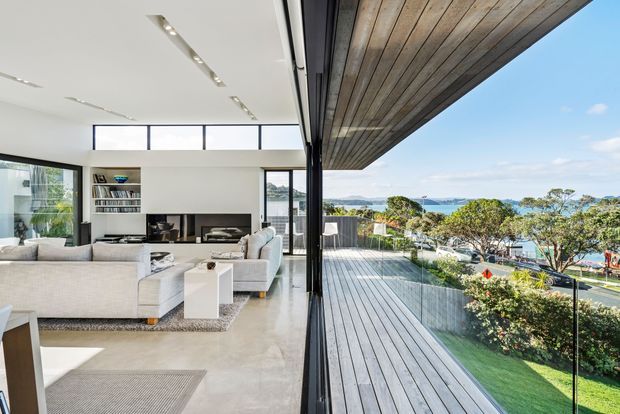 Bright contemporary open-plan living room opening onto a large timber deck through full-height glazed doors. The interior features pale upholstered sofas, a built-in dark fireplace and media wall, white cabinetry, polished concrete flooring, and linear recessed lighting. The deck has a broad timber soffit and glass balustrade, with landscaped greenery and a visible coastal water view beyond.
