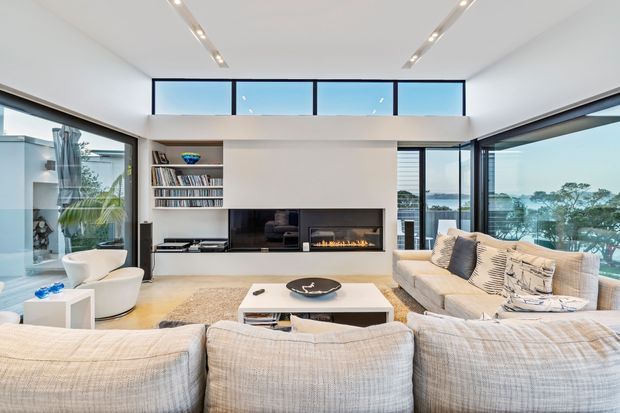 Bright contemporary living room with expansive floor-to-ceiling glazing and high clerestory windows overlooking a leafy waterfront or coastal setting. A large pale sectional sofa faces a built-in media wall with open shelving, television, and linear fireplace. The room also includes a white lounge chair, pale rug, low rectangular coffee table, recessed linear ceiling lights, and light neutral finishes.