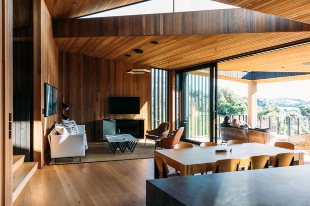 Open-plan contemporary living, dining, and kitchen interior with extensive dark timber wall and ceiling lining, warm timber flooring, and large glazed openings connecting to a covered outdoor terrace. The living area includes a fireplace, television, sofa, and leather lounge chairs, while a substantial timber dining table sits beside a dark stone kitchen island. Strong indoor-outdoor connection and views beyond the terrace are visible.