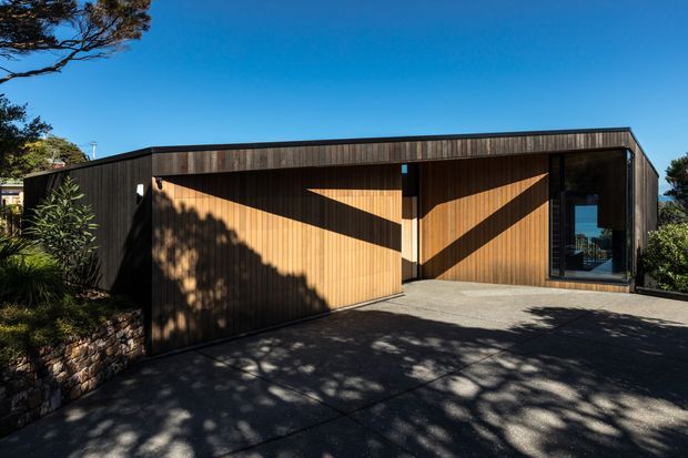 A low, modern single-storey house viewed from the driveway, with a dark timber-clad facade, broad projecting roofline, and a large timber garage door featuring strong geometric sunlight and shadow patterns. A glazed entry or living-space opening is visible at right, with landscaped planting and stone retaining walls around the paved driveway.