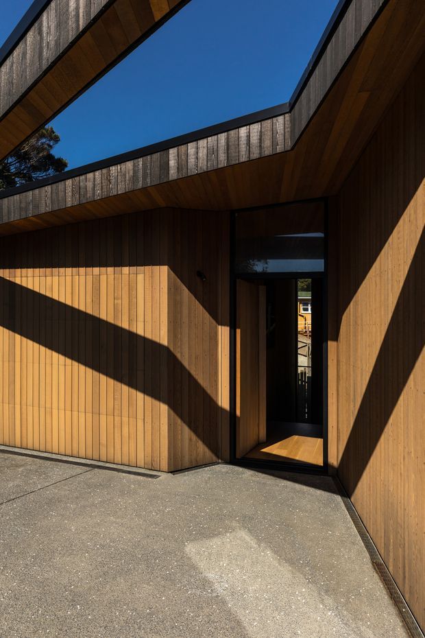 A contemporary timber-clad residential entrance or covered courtyard viewed from outside. Vertical warm-toned wood boarding wraps the walls and soffits, with dark metal-framed glazing and door openings. Angular roof planes and clerestory-like openings frame a clear blue sky, while strong sunlight creates dramatic geometric shadows across the facade and concrete paving.