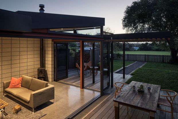 A contemporary indoor-outdoor living space with a low, dark flat-roofed extension opening onto a timber deck and grassed yard. The room features large glazed sliding doors, warm timber-framed glazing, a pale tiled floor, tan sofa seating, and a compact freestanding fireplace. A timber outdoor dining table occupies the foreground deck, while the garden and mature tree are visible beyond.