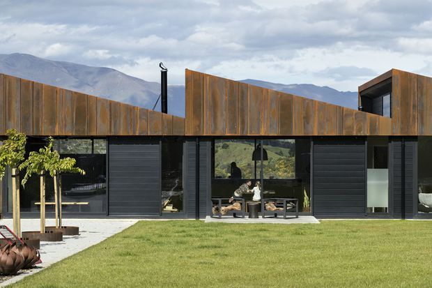 Single-storey contemporary house viewed from the lawn, with a long low facade combining dark glazing and garage-style doors beneath a sculptural weathered metal roof and parapet. The oxidised brown metal cladding forms angular folded rooflines, while the black-framed glazing reflects the surrounding mountainous landscape. A paved terrace, outdoor seating, lawn, and young trees occupy the foreground.