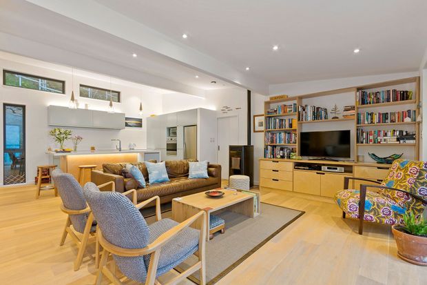 Bright open-plan living and dining area with a contemporary white kitchen, timber flooring, built-in timber media and bookcase joinery, a central sofa, dining table and patterned armchairs. Recessed ceiling lighting and clerestory windows create an airy, welcoming interior.