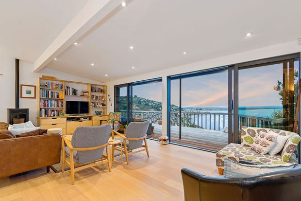 A bright, open-plan living room with warm timber flooring, built-in timber shelving and media cabinetry, a large seating arrangement, and extensive sliding glass doors opening to a balcony with a coastal outlook. Recessed ceiling spotlights and contemporary glazing create an airy, relaxed interior.