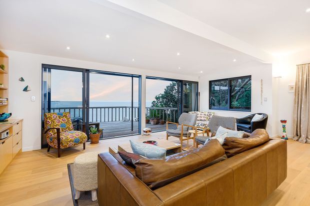 Bright, open-plan living room with warm timber flooring, a large brown leather sectional sofa, patterned occasional chairs, a light coffee table, and extensive black-framed glazing. The glazing opens onto a balcony with a railing and expansive coastal or ocean views. Built-in timber shelving and cabinetry line the left side, while recessed ceiling lights and neutral walls create a relaxed contemporary interior.