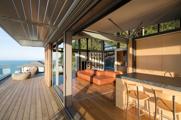 A bright, open-plan coastal living and kitchen space opening onto a timber-clad outdoor deck. Full-height sliding glass doors create a seamless indoor–outdoor connection, with ocean views beyond. Inside, the room features warm timber flooring and cabinetry, a dark stone kitchen benchtop with integrated seating, a low rust-orange leather sectional, and a compact bar or display joinery area. High-level glazing and exposed structural elements enhance the airy, contemporary feel.