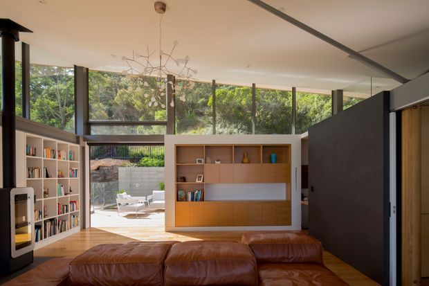 A warm, mid-century-influenced living room with a large brown leather sectional in the foreground, timber flooring, built-in timber shelving and cabinetry, and extensive floor-to-ceiling glazing opening toward a landscaped outdoor terrace. A sculptural pendant light and a dark partition wall add architectural interest, while the room feels connected to the surrounding greenery.
