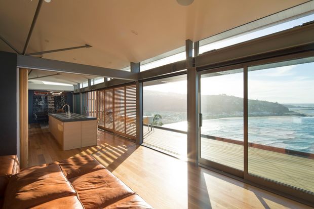 A warm, contemporary open-plan interior overlooking the ocean, with a kitchen island, timber flooring, expansive sliding glass doors, timber sun-shading screens, and a low brown leather sofa in the foreground. Strong sunlight creates striped shadows across the floor, while the glazed facade opens to a deck and coastal view.