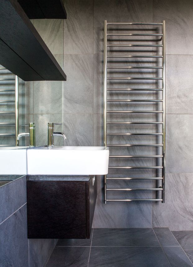 A compact contemporary bathroom featuring large grey stone-look wall and floor tiles, a wall-mounted white rectangular basin, dark timber vanity cabinetry, chrome tapware, a large frameless mirror, and a vertical heated towel rail. The palette is monochromatic and refined with strong geometric lines.