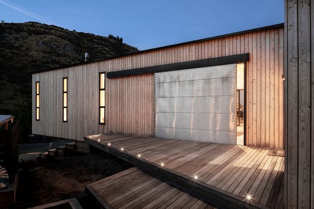 Exterior view of a modern, elongated building clad in vertical timber boards, set against a rugged hillside. A broad timber deck and stepped walkway with integrated spot lighting lead to a large metallic sliding or folding door, with narrow vertical windows punctuating the facade.