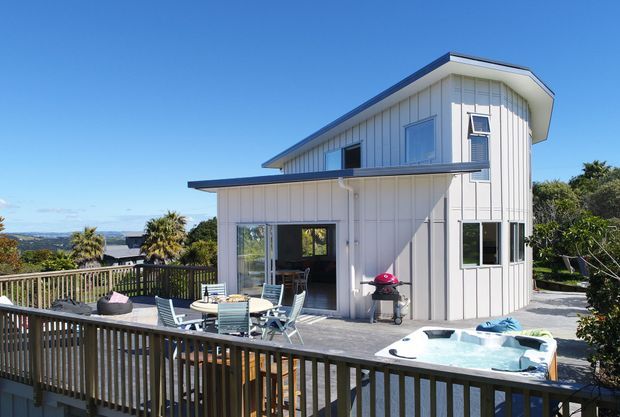 Exterior view of a contemporary two-storey white-clad house with a distinctive sloping roofline, broad timber deck, outdoor dining furniture, and a spa pool. The elevated deck overlooks a leafy coastal or suburban landscape under a clear blue sky.