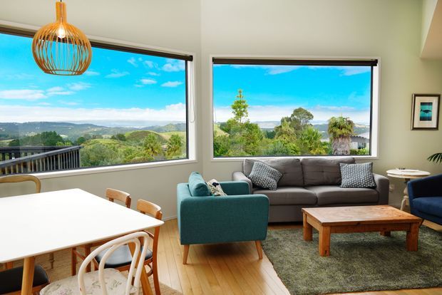 Bright open-plan living and dining area with large picture windows overlooking a green, hilly landscape and water. The room features pale timber flooring, light walls, a white dining table with curved chairs, a teal armchair, a charcoal sofa, a rustic timber coffee table, and a woven pendant light.