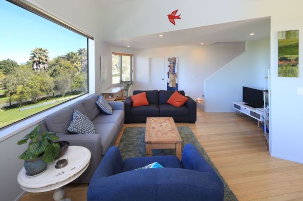 Bright contemporary living room with expansive corner glazing overlooking landscaped greenery. The room features pale timber flooring, white walls, a grey sectional sofa, navy armchairs, orange cushions, a timber coffee table, and a compact TV unit. A lowered ceiling zone with recessed lighting and a red bird wall feature add visual interest.