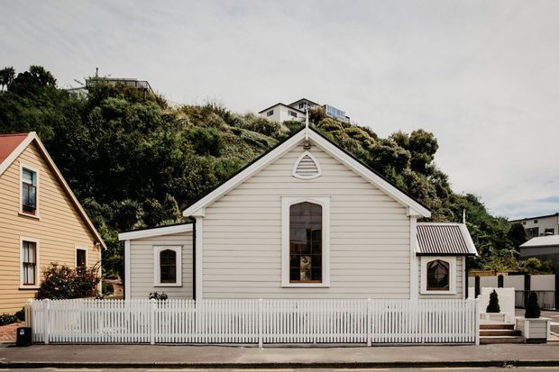 Street-facing view of a compact white weatherboard cottage with a steep gabled roof, central arched-top window, decorative gable vent, and symmetrical side extensions. A white picket fence encloses the frontage, with neighbouring timber homes and a densely vegetated hillside behind.
