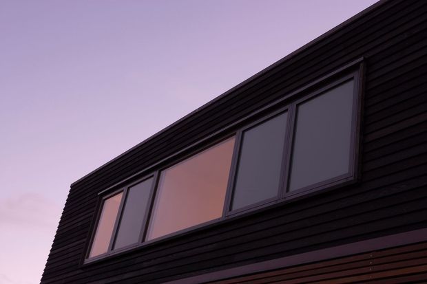 Close-up architectural exterior view of a dark, horizontally clad upper facade with a long band of modern windows. The glazing reflects a soft pink-purple evening sky, while horizontal timber-like cladding and clean linear framing create a dramatic contemporary appearance.