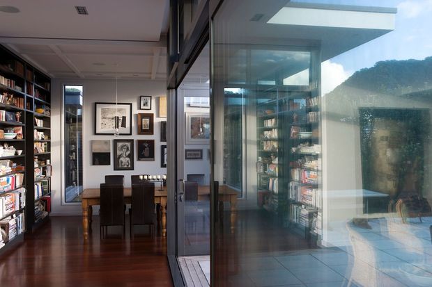 Interior view of a compact contemporary library and dining area, with floor-to-ceiling glazing creating strong reflections and connecting the dark timber-floored room to a bright tiled outdoor or glazed extension. Full-height bookshelves line the left wall, while a timber dining table, dark upholstered chairs, framed artwork, and extensive glass partitions define the space.