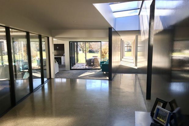 A bright, open-plan contemporary interior with polished concrete flooring, black-framed full-height glazing and internal glass partitions. A long skylight brings daylight into the space, while a deep black feature wall and framed photographs add contrast. The room opens toward a small outdoor terrace and garden beyond.