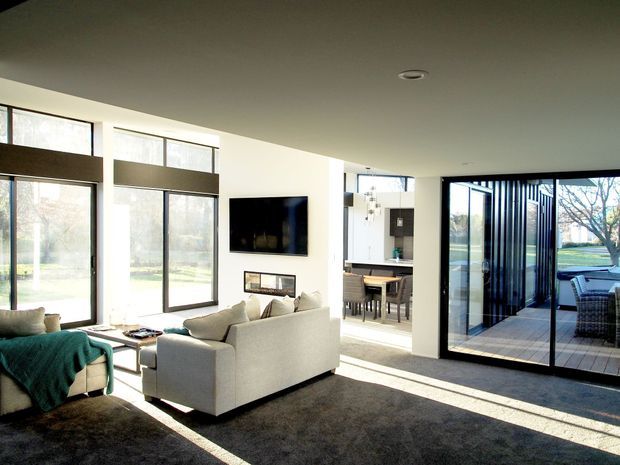 Bright contemporary living room with large floor-to-ceiling windows and expansive black-framed glazed doors opening to a timber deck. The room contains light upholstered sofas, a dark area rug, wall-mounted television, and a dining and kitchen area visible beyond. Strong indoor-outdoor connection and abundant natural light.