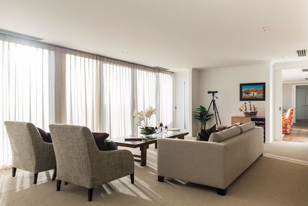 Bright, spacious contemporary living room with full-height sheer curtains, neutral upholstered seating, a low timber coffee table, and a beige sofa arranged around the lounge area. A hallway and adjoining rooms are visible beyond the living space, with decorative artwork and a tripod adding visual detail.