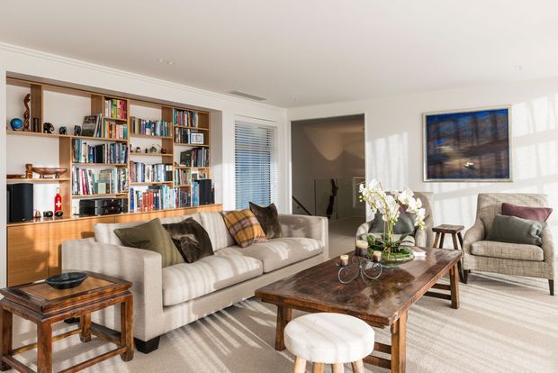 Bright, elegant living room with a large built-in timber bookcase and cabinetry, upholstered sofa and armchair seating, a substantial dark wood coffee table, and framed artwork. Strong daylight enters through the window, creating shadows across the neutral carpet and furniture.