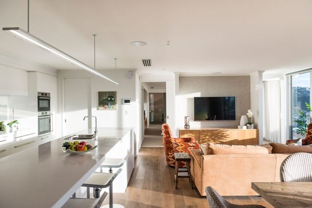 Bright open-plan contemporary living space combining a white kitchen with a long stone island, integrated appliances and slim linear pendant lighting, opening to a warm living and dining area. The living room features a tan sectional sofa, timber media console, wall-mounted television and generous glazing with vertical blinds. Timber flooring and neutral finishes create a spacious, welcoming interior.