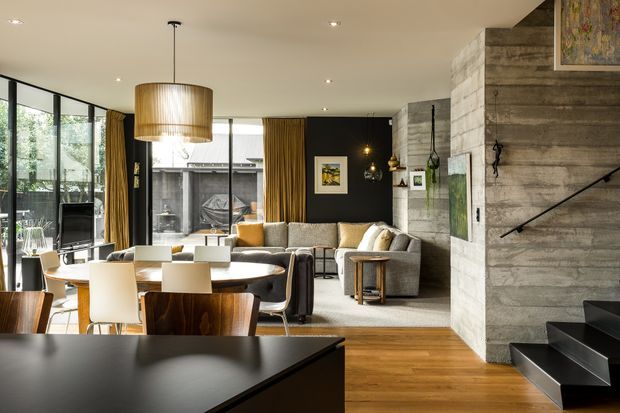 Open-plan contemporary living and dining area with a partial kitchen island in the foreground. The room features floor-to-ceiling glazing, warm timber flooring, a large pendant light over the dining table, neutral upholstered seating, dark feature walls, and a prominent textured concrete or stone stair enclosure with black floating stairs and a timber sliding door.