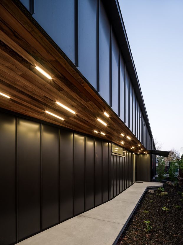 A dramatic dusk exterior view of a long, contemporary building facade. The lower level is clad in dark vertically panelled metal, while the upper facade features tall dark-framed vertical glazing or panels. A warm timber soffit with integrated linear lighting projects over a narrow concrete walkway, bordered by landscaped planting beds.