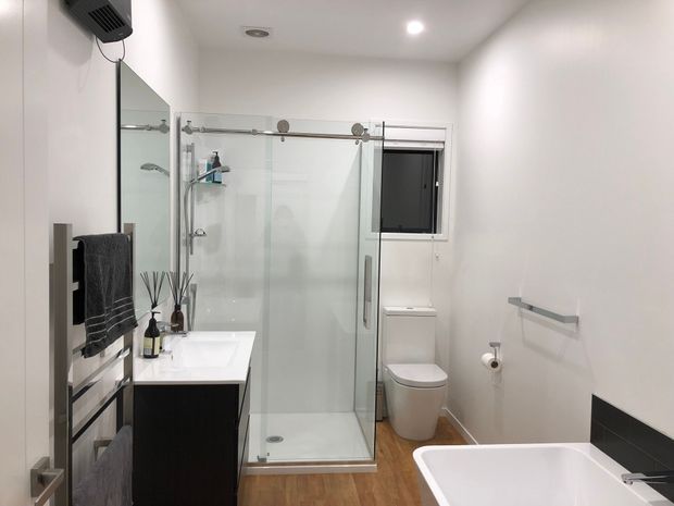 Bright contemporary bathroom featuring a frameless glass walk-in shower with exposed sliding hardware, a dark floating vanity with white basin, toilet, freestanding white bathtub, heated towel rail, and warm wood-look flooring. The room has white walls, recessed lighting, and a small dark-framed window.