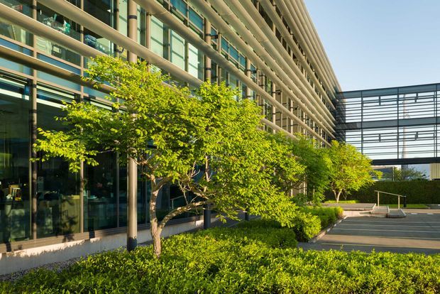 Exterior view of a contemporary multi-storey commercial building with extensive glass curtain walls, horizontal external sun-shading fins, and a landscaped forecourt with small trees and clipped hedges. A paved access or parking area and a glazed connecting structure are visible to the right.