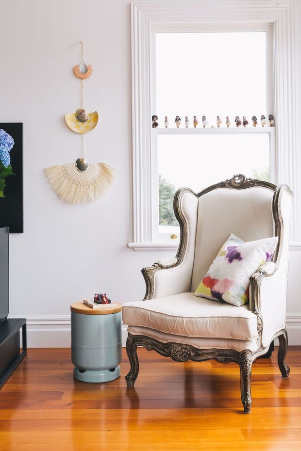 Bright, eclectic sitting area featuring an ornate upholstered wingback armchair with carved dark wood frame, a small cylindrical blue side table, polished timber flooring, white walls, and a large white-trimmed window. Decorative woven wall hanging and a small row of figurines add playful character, while a partial television is visible at left.