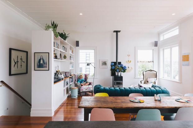 Bright, open-plan living and dining area with white walls, built-in open shelving, polished timber flooring, and large windows. A deep teal sofa faces a compact black fireplace with a vertical flue, while a rustic timber dining table with colourful upholstered chairs occupies the foreground. The room includes framed artwork, bookshelves, indoor plants, and a stairway descending at the left.