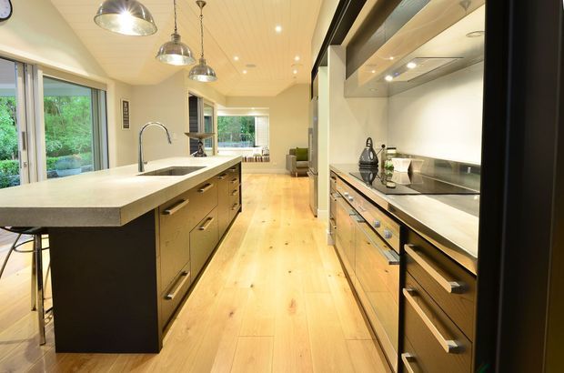 A contemporary galley-style kitchen with a large central island, integrated sink and tall curved faucet, extensive timber cabinetry, pale engineered-looking benchtops, stainless-steel appliances, and warm timber flooring. Pendant lights hang above the island, while large windows provide views to greenery and bring in natural light.
