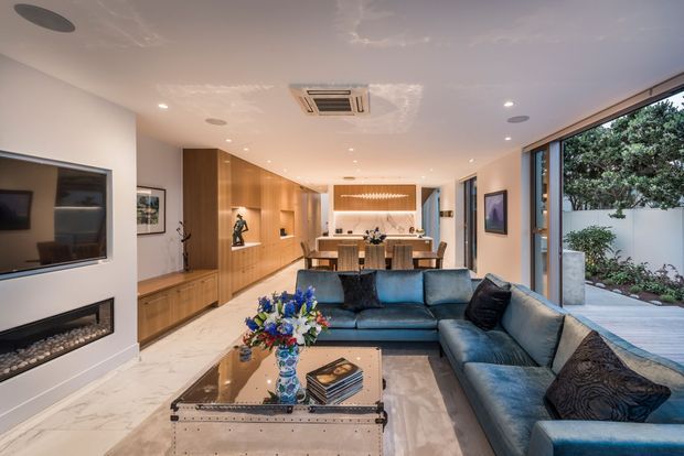 Contemporary open-plan living room with a large blue sectional sofa, marble-look flooring, a mirrored metal coffee table, built-in timber cabinetry and display shelving, a wall-mounted television, and a linear fireplace. The room opens through wide glazing to a landscaped outdoor area and includes a dining and kitchen zone at the rear. Recessed lighting and concealed air conditioning create a polished, refined interior.