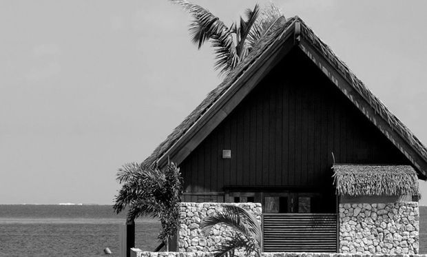 Black-and-white exterior view of a tropical, steeply pitched gable-roof building beside the sea. The structure has a thatched roof and thatched awning, dark vertical timber wall cladding, and rough light-colored stone walls, with palm trees visible behind and the ocean in the background.