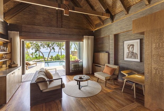 A warm, richly textured living room with a high exposed timber-beam and woven ceiling, dark timber wall cladding, and timber flooring. The room contains a sofa, lounge chairs, round coffee table, rug, built-in shelving and a compact desk. Large sliding glass doors open to a timber deck, swimming pool and lush coastal landscape beyond. Decorative patterned timber panels and framed artwork add visual interest.