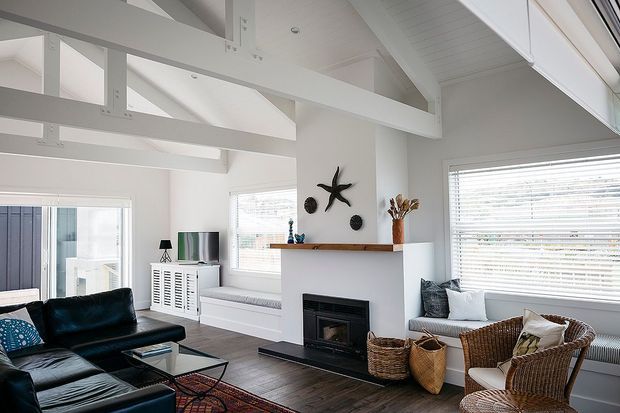 Bright coastal-inspired living room with a high pitched ceiling and exposed white-painted structural beams. The room features a dark leather sectional, woven rattan armchairs and baskets, a central white fireplace wall with timber mantel, large windows with white blinds, and dark timber flooring.