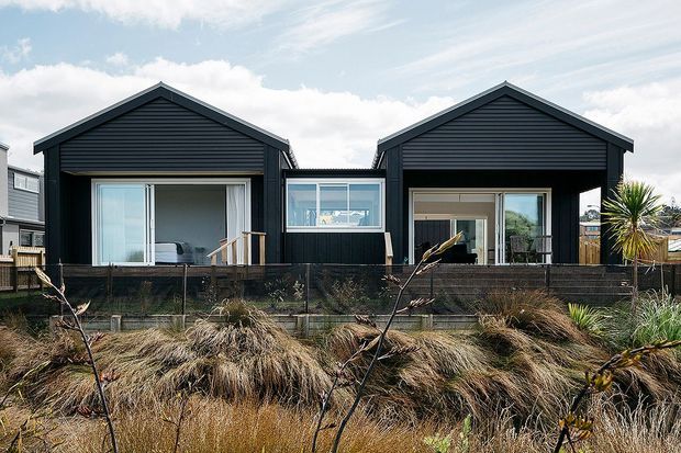 Exterior view of a contemporary single-storey residential building composed of two dark-clad gabled volumes connected by a central glazed section. Large sliding glass doors open onto a raised timber deck, with coastal-style grasses and planting in the foreground.