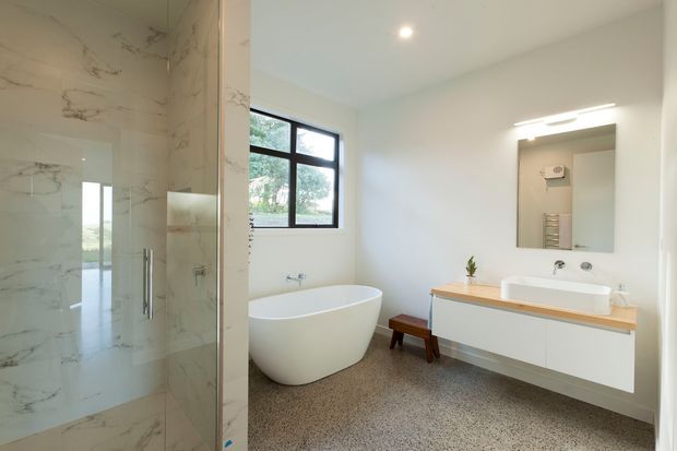 Bright contemporary bathroom featuring a freestanding white bathtub, frameless glass shower enclosure with marble-look wall tiles, floating white vanity with a timber benchtop, large mirror, and black-framed window. The room has a clean, refined, spa-like character.