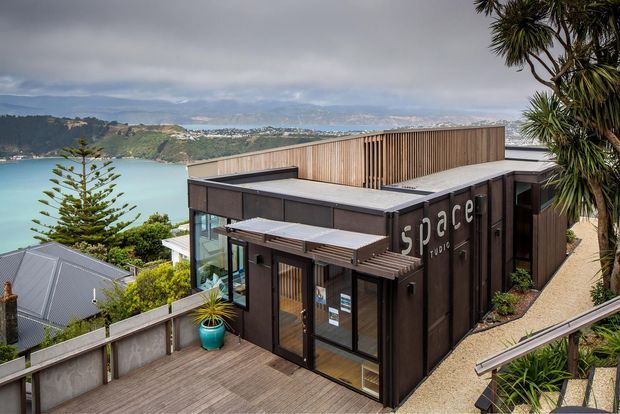 Exterior view of a compact modern multi-level building with dark vertical cladding, timber screening, flat roofs, glazed entry doors, and a timber deck. The building sits on a steep coastal hillside overlooking a sheltered bay, with neighbouring homes and subtropical planting visible around it. The facade prominently displays the word “space,” suggesting a commercial or studio premises.