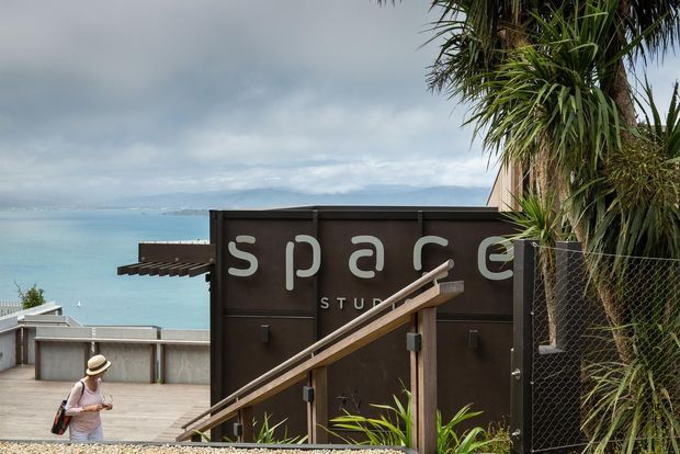 Exterior view of a dark-clad studio building beside a turquoise waterfront, with a prominent “space studio” sign, timber handrail and steps, concrete terrace, and coastal planting including tall palm-like vegetation. A person is visible on the lower terrace, while the building and waterfront setting dominate the frame.