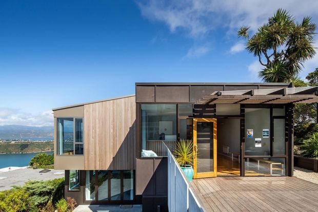 Exterior view of a modern multi-level home on a sloped waterfront site. The house combines vertical timber cladding, dark metal panels, extensive glazing and a broad timber deck, with a covered entry and pergola-like roof structure. Water and distant hills are visible beyond the property.