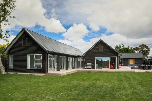 Exterior view of a single-storey dark-clad residential house with multiple pitched gable roofs, extensive glazing, timber decking, and a broad manicured lawn. The composition shows a main gabled wing connected to a lower central link and a second gabled wing, with an outdoor entertaining area visible to the right.