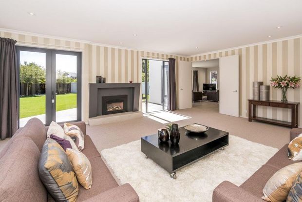 Bright, spacious living room with a contemporary fireplace, large black-framed glazed doors opening to a lawn, beige carpet, a large cream rug, and two taupe sofas arranged around a black coffee table. Neutral striped wallpaper and recessed ceiling lighting create a refined, traditional-contemporary interior.