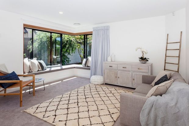 Bright, airy living room with a bay-style corner window seat, floor-to-ceiling glazing, white walls, pale carpet, a textured neutral rug, a large sectional sofa, timber-framed lounge chair, and a light painted timber sideboard. Sheer curtains and lush greenery visible outside create a calm indoor-outdoor feel.